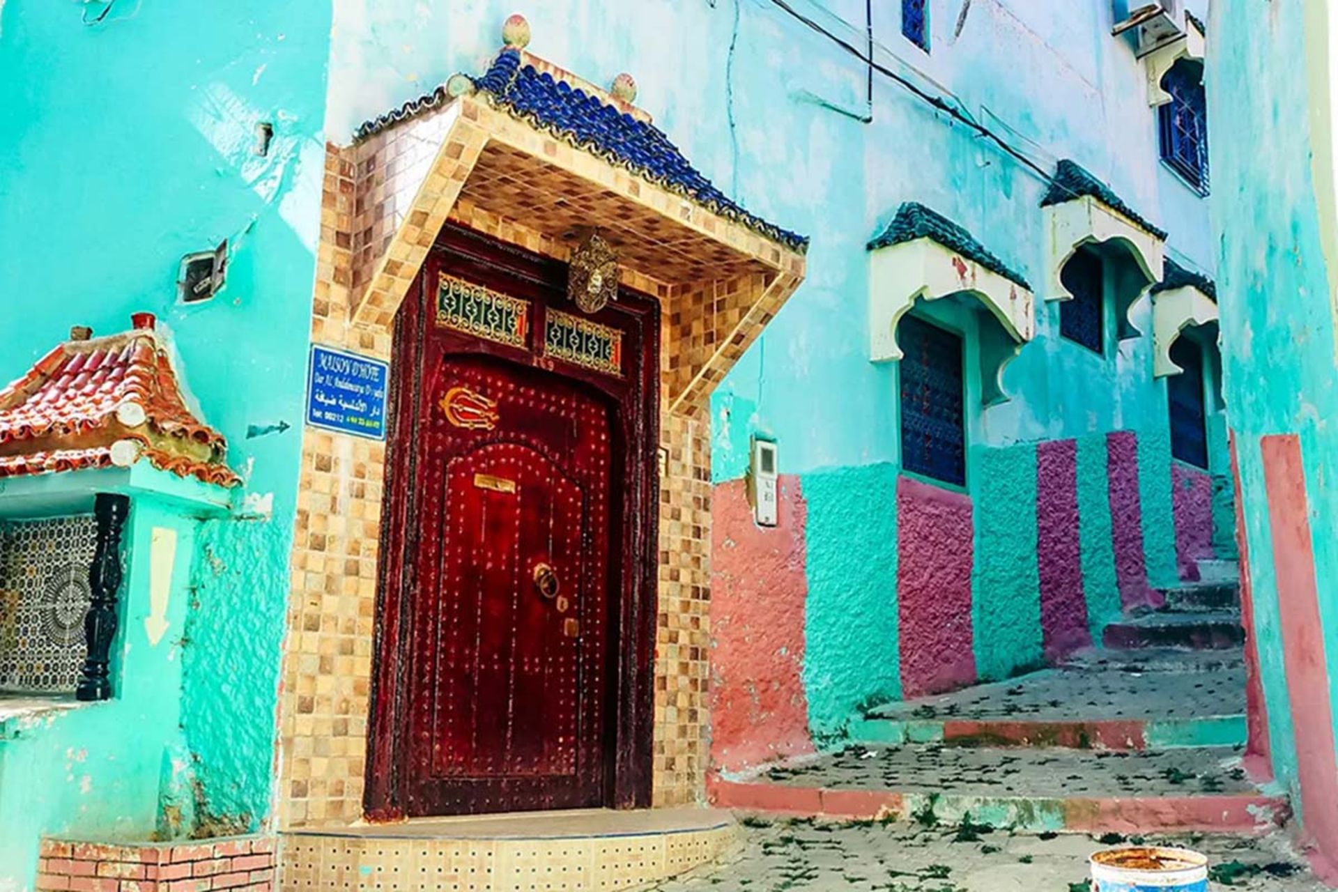 نمای خانه‌ای رنگارنگ در مولای ادریس با درب چوبی قرمز تیره و دیوارهای فیروزه‌ای و مرجانی