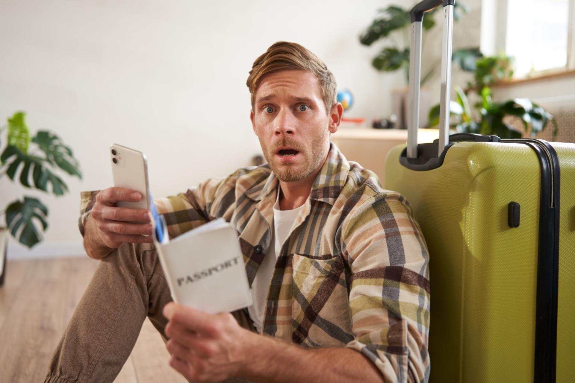 فردی متعجب با در دست داشتن پاسپورت و تلفن همراه، تکیه داده به چمدان سبز